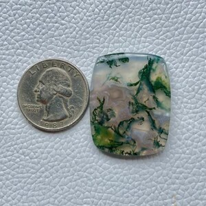 Pode incluir: Uma pedra preciosa de ágata musgosa retangular com inclusões verdes, brancas e transparentes, ao lado de uma moeda de um quarto de dólar americana. A pedra preciosa tem cantos arredondados e uma superfície polida.