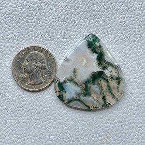 Pode incluir: Uma pedra preciosa de ágata musgo em forma de gota com padrões verdes, brancos e castanhos. Uma moeda de um quarto de dólar dos EUA está ao lado da gema para comparação de tamanho. A gema está sobre uma superfície texturizada cinza claro.