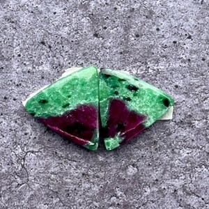 May include: Two triangular gemstones, each featuring a vivid green upper section and a rich burgundy base. The green area is speckled with black. The gemstones are displayed on small white paper pieces, highlighting their natural colours and patterns.