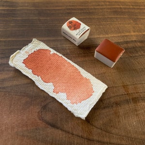 May include: A small, square watercolour pan with a red-brown pigment. The pan is labelled "Red Clover Meadow" and "England Red". A sample of the watercolour paint is shown on a piece of paper.