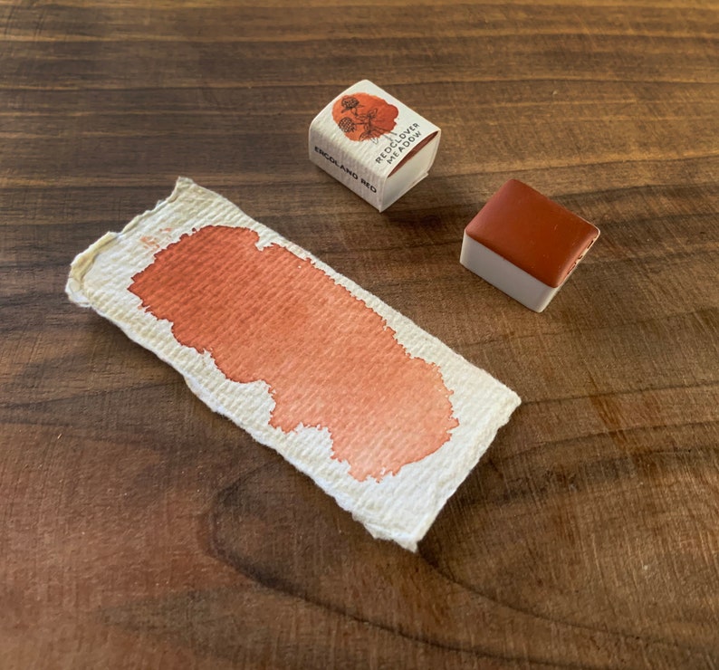 May include: A small, square watercolour pan with a red-brown pigment. The pan is labelled "Red Clover Meadow" and "England Red". A sample of the watercolour paint is shown on a piece of paper.