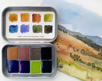Puede incluir: Una lata de metal con un juego de 8 acuarelas. La lata está abierta y las pinturas están dispuestas en dos filas de cuatro. Los colores son marrón, naranja, verde, azul, morado y negro. La lata tiene el sitio web www.redclovermeadow.com impreso en la parte delantera.