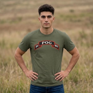 May include: An olive green short-sleeve crew neck t-shirt featuring a graphic design on the chest. The graphic is a black banner with a red border containing the white text 1st POG RGT, arranged in an arched formation across the front of the shirt.