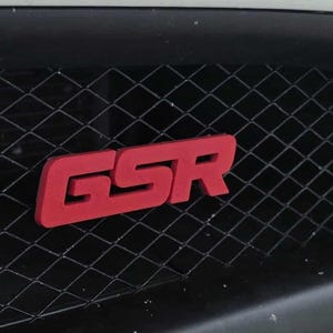 May include: Close-up of a black vehicle grille with a red "GSR" emblem. The emblem is a bold, three-dimensional design, and the grille has a diamond-shaped pattern. The vehicle's bumper is visible.