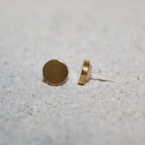 May include: Two small, round, gold-colored stud earrings. One earring is positioned upright, while the other is on its side, revealing the post. The earrings are set against a textured, light gray surface.