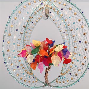 Op de afbeelding: Een unieke hanglamp met een lichtblauw draadframe, versierd met gouden sterren en kralen. Binnenin staat een kleurrijke boom met levendige bladeren in rood, oranje, geel en roze, wat een speels tintje geeft.