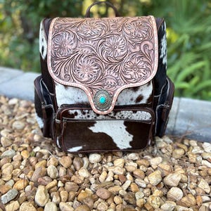 May include: A backpack featuring a brown and white cowhide pattern with a tan leather flap embossed with a floral pattern. The flap is secured by a silver-toned concho ornament with a central turquoise stone. It includes a front zippered pocket.