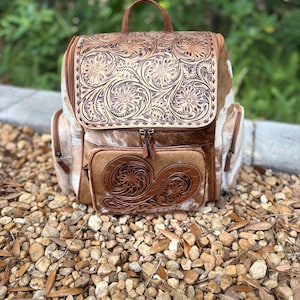 May include: A brown and tan backpack with a floral tooled leather flap and pocket. The backpack has a handle and two side pockets. The body of the bag is made of cowhide with white and brown patches. The bag is sitting on a bed of small rocks.