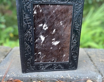 May include: A rectangular notebook cover featuring a central panel of brown and white cowhide hair-on leather, framed by a wide black leather border with intricate, embossed floral scrollwork patterns along the edges.