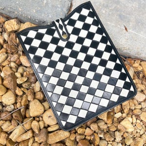 May include: A black and white checkered pattern tablet case with a leather border and a snap closure. The case is rectangular and rests on a bed of small rocks and pebbles.