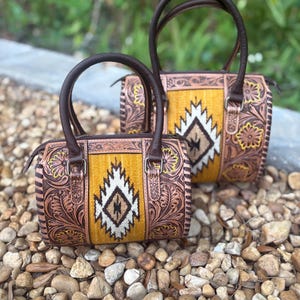 May include: Two brown leather barrel bags with intricate floral and geometric designs. The bags feature a mustard yellow center panel with a white, brown, and black pattern. The bags have dark brown handles and trim, and are set on a bed of small rocks.