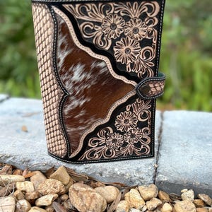 May include: A handcrafted leather journal with a unique design. The cover features a combination of textured tan leather, brown and white cowhide, and intricate floral tooling. A black border and a snap closure complete the design. The journal is a stylish accessory.
