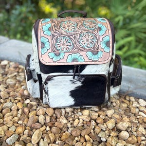 May include: A backpack featuring a black and white cowhide body with brown leather trim. The top flap displays a tan leather panel with intricate floral tooling and hand-painted turquoise accents on the flower petals and surrounding scrollwork.