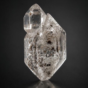 May include: A clear, double-terminated quartz crystal cluster featuring a smaller crystal attached to the side of a larger, hexagonal prism. The mineral specimen contains visible dark brown and black inclusions scattered throughout its structure.