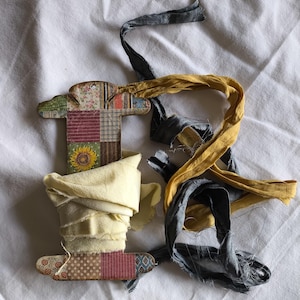 May include: A wooden spool with a patchwork design, holding a roll of pale yellow fabric. Grey and gold ribbons are draped around the spool. The spool has a sunflower and floral pattern.