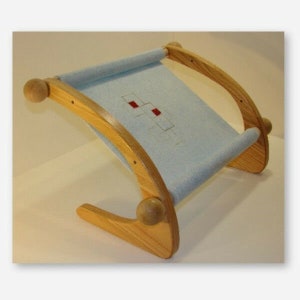 Könnte beinhalten: Ein Holzständer für einen Stickrahmen mit einem hellblauen Stoff, der über den Rahmen gespannt ist. Der Ständer hat eine gebogene Basis und ist aus Naturholz gefertigt.