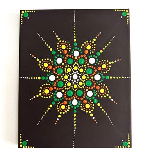 Op de afbeelding: Een zwart canvas met een kleurrijk stippenmandala-ontwerp. De mandala is gemaakt van gele, oranje, groene en witte stippen. Het ontwerp is symmetrisch en heeft een stervorm.
