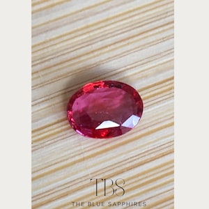 May include: A vibrant, oval-cut, red gemstone displayed on a light brown, textured surface. The image includes the text "TBS THE BLUE SAPPHIRES" at the bottom.