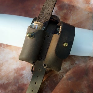 K&ouml;nnte beinhalten: Ein brauner Lederg&uuml;rtel mit zwei kleinen Glasfl&auml;schchen, die daran befestigt sind. Die Fl&auml;schchen haben Korkstopfen und werden durch Lederriemen gehalten. Der G&uuml;rtel hat eine Schnalle und ist mit N&auml;hten verziert.