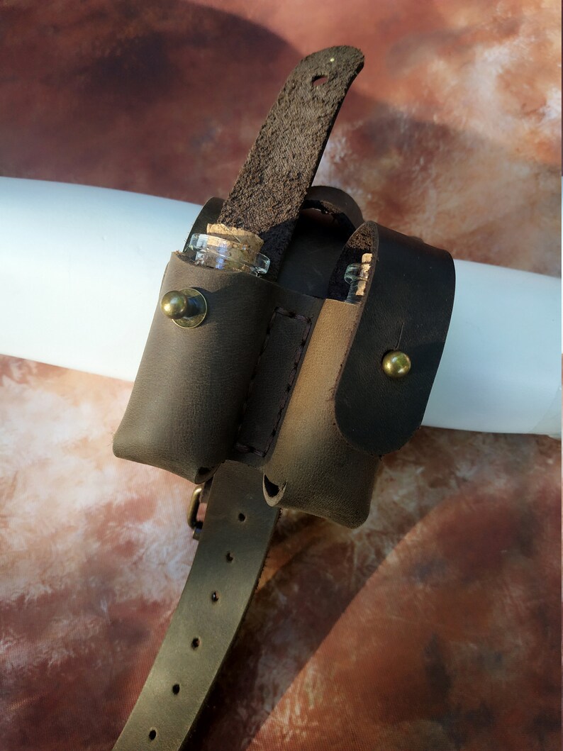 K&ouml;nnte beinhalten: Ein brauner Lederg&uuml;rtel mit zwei kleinen Glasfl&auml;schchen, die daran befestigt sind. Die Fl&auml;schchen haben Korkstopfen und werden durch Lederriemen gehalten. Der G&uuml;rtel hat eine Schnalle und ist mit N&auml;hten verziert.