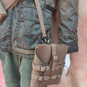 May include: A brown leather book bag with multiple buckles and straps. The bag is worn over the shoulder and has a strap that goes across the chest.