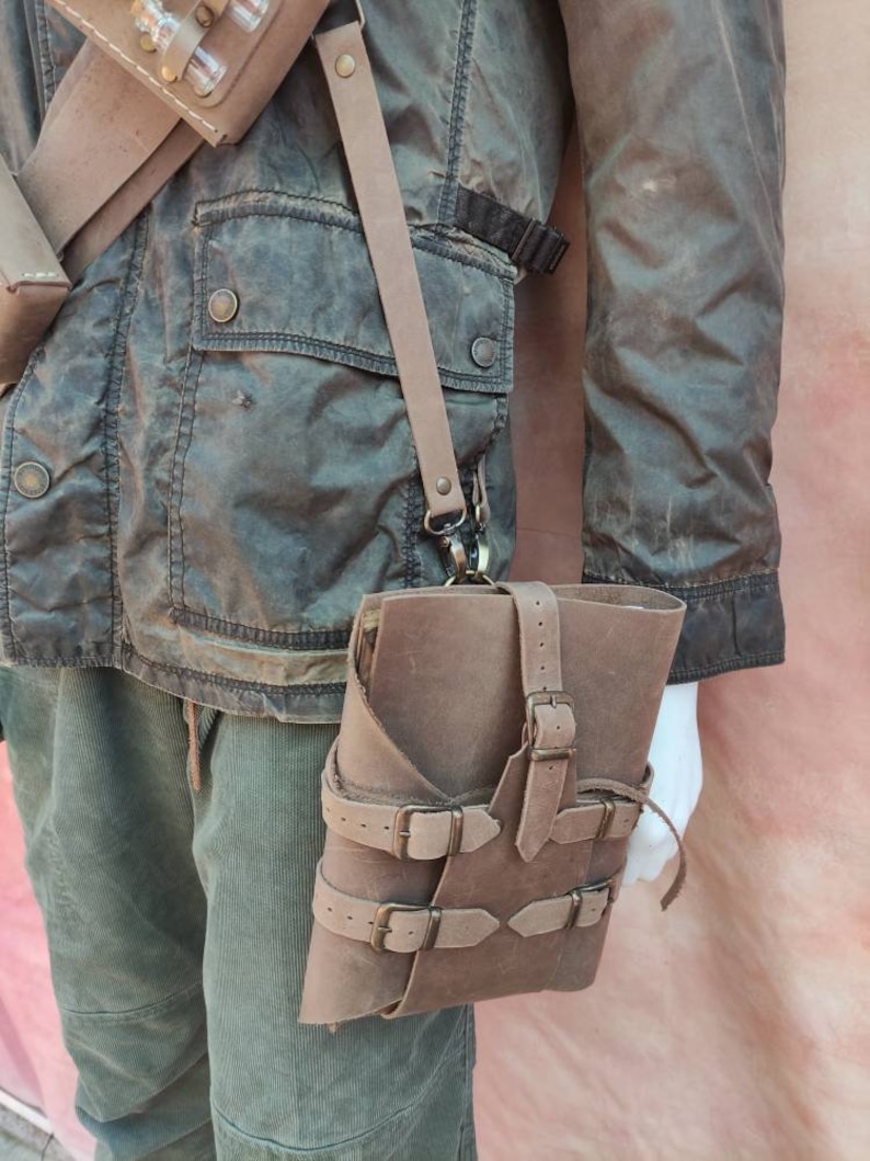 May include: A brown leather book bag with multiple buckles and straps. The bag is worn over the shoulder and has a strap that goes across the chest.