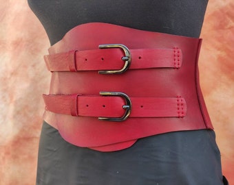 May include: A red leather corset with two double buckles. The corset has a smooth, shiny finish and is designed to cinch the waist.