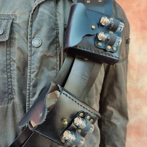 May include: A black leather shoulder holster with two pouches, each holding three small glass vials with cork stoppers. The holster is worn over a brown jacket.