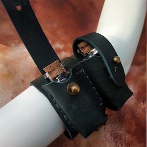 May include: A dark green leather wrist cuff with two small glass vials with cork stoppers. The vials are held in place by leather straps and secured with metal snaps.