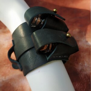 K&ouml;nnte beinhalten: Ein dunkelgr&uuml;nes Lederarmband mit zwei Metallringen und Messingakzenten. Das Armband ist so konzipiert, dass es kleine Gegenst&auml;nde wie Schl&uuml;ssel oder ein Messer aufnehmen kann.