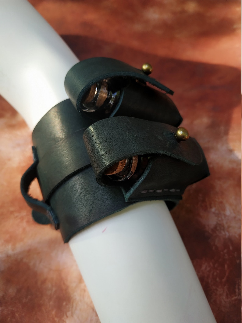 K&ouml;nnte beinhalten: Ein dunkelgr&uuml;nes Lederarmband mit zwei Metallringen und Messingakzenten. Das Armband ist so konzipiert, dass es kleine Gegenst&auml;nde wie Schl&uuml;ssel oder ein Messer aufnehmen kann.
