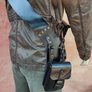 May include: A black leather thigh holster with a strap and buckle. The holster has a flap with a buckle and is decorated with brass studs.
