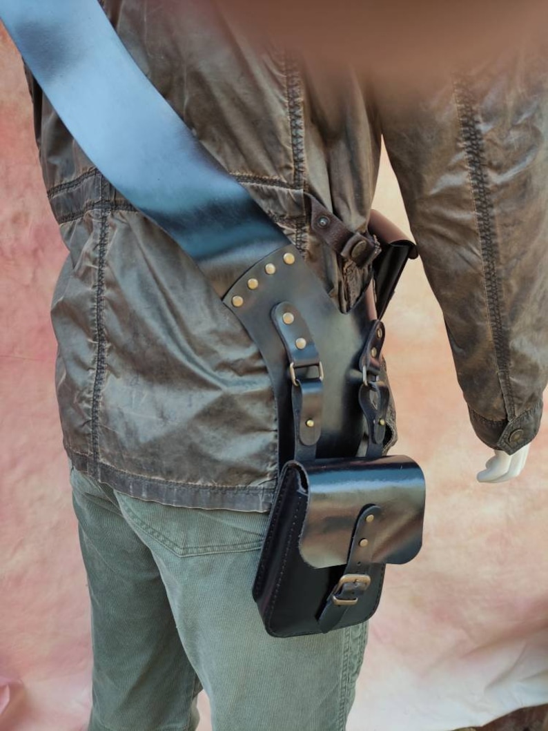 May include: A black leather thigh holster with a strap and buckle. The holster has a flap with a buckle and is decorated with brass studs.