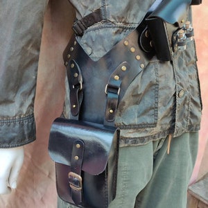 May include: A black leather belt with multiple pouches and straps. The belt has a Y-shaped design and is decorated with brass studs. The belt is worn over a brown jacket and green pants.