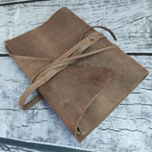 May include: A brown leather journal with a strap closure. The journal is closed and lying on a wooden surface.