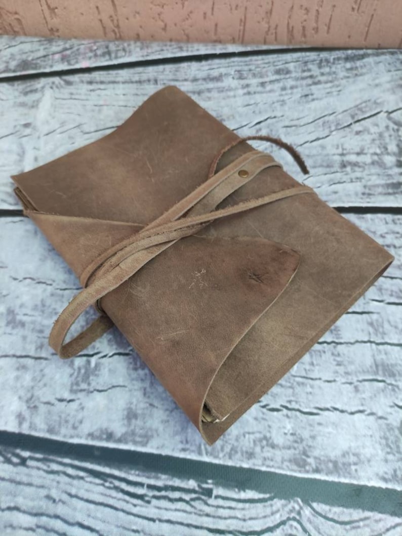 May include: A brown leather journal with a strap closure. The journal is closed and lying on a wooden surface.