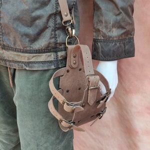 May include: A brown leather holster with multiple buckles and straps. The holster is attached to a brown leather strap with a metal clasp.
