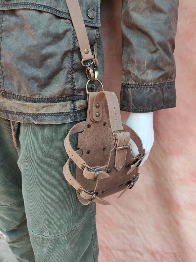May include: A brown leather holster with multiple buckles and straps. The holster is attached to a brown leather strap with a metal clasp.