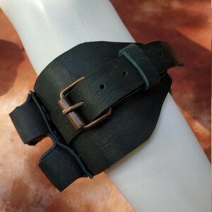 K&ouml;nnte beinhalten: Ein dunkelgr&uuml;nes Lederarmband mit einer Messing-Schnalle. Das Band ist verstellbar und hat einen Riemen, der sich um den Arm wickelt.