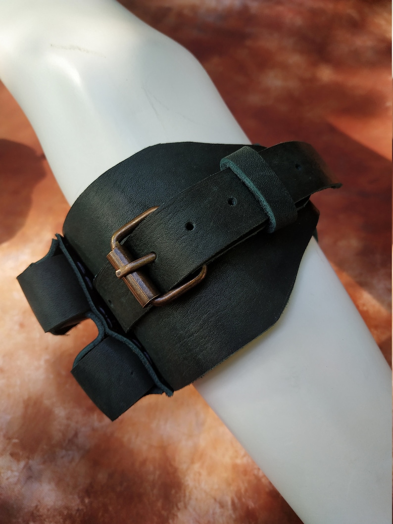 K&ouml;nnte beinhalten: Ein dunkelgr&uuml;nes Lederarmband mit einer Messing-Schnalle. Das Band ist verstellbar und hat einen Riemen, der sich um den Arm wickelt.
