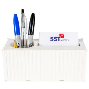 May include: A white desk tidy in the shape of a shipping container, containing a blue marker, a black marker, a pen, a pencil, and a business card with the text "SST MODELS". The organiser has three compartments.