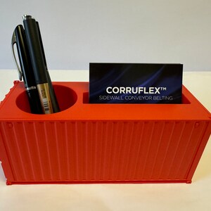Könnte beinhalten: Ein roter Plastikbehälter in Form eines Versandcontainers mit zwei Fächern für Stifte und einem Fach für Visitenkarten. Das Fach für Visitenkarten enthält eine Visitenkarte mit dem Text "CORRUFLEX™ SIDEWALL CONVEYOR BELTING".