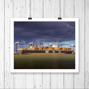 Może przedstawiać: Duży statek towarowy z wieloma kontenerami ułożonymi na pokładzie. Statek jest zacumowany w porcie z żurawiami w tle. Statek jest oznaczony jako "M.S.C."