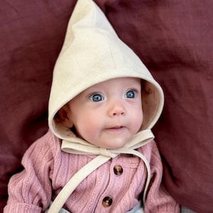 May include: A baby wearing a light beige bonnet with a pointed top and a pink knitted sweater. The bonnet has a long tie that is tied under the chin.