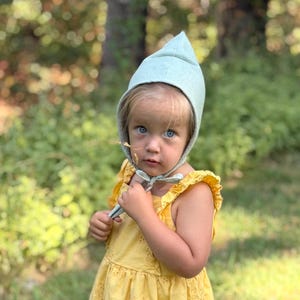 May include: A light green pointed bonnet with a chin tie, paired with a yellow sleeveless dress featuring eyelet embroidery and ruffled shoulder straps. The bonnet has a structured, peaked top and covers the head, while the dress has a gathered skirt.