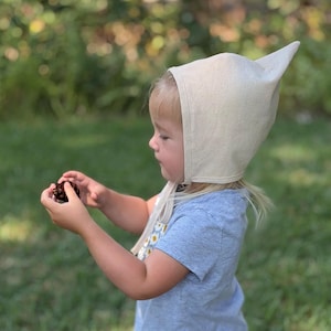 May include: A cream-colored pointed bonnet made of fabric with a tie closure under the chin, worn with a light blue short-sleeved t-shirt featuring a small floral pattern on the front, while holding a small brown pinecone in the hands.