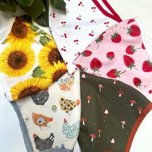May include: A collection of five fabric face masks with various patterns. Designs include sunflowers, cherries, strawberries, chickens, and mushrooms. Each mask has a different trim color, and the masks are arranged on a white surface.