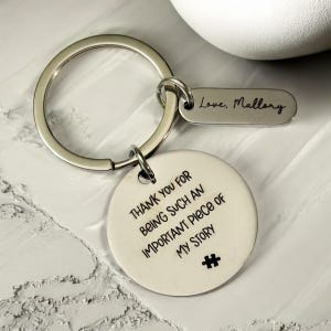 May include: A silver-colored keychain with a round pendant and a smaller rectangular tag. The round pendant has the text "THANK YOU FOR BEING SUCH AN IMPORTANT PIECE OF MY STORY" and a puzzle piece graphic. The rectangular tag reads "Love, Mallory".