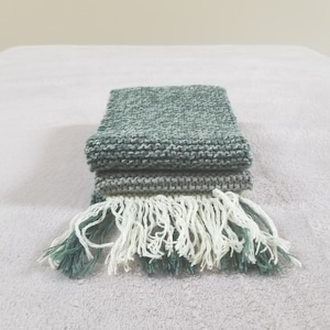 May include: A folded green knitted scarf with white fringe. The scarf is made of a soft, textured yarn.