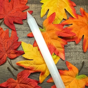 Puede incluir: Una varita de selenita blanca se encuentra en diagonal sobre una superficie de madera, rodeada de coloridas hojas de oto&ntilde;o en tonos rojos, naranjas y amarillos. La varita es cil&iacute;ndrica con una punta. Las hojas a&ntilde;aden un toque estacional.